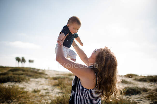 Grand-mère tenant petit-fils haut tout en se tenant sur la plage — Photo de stock