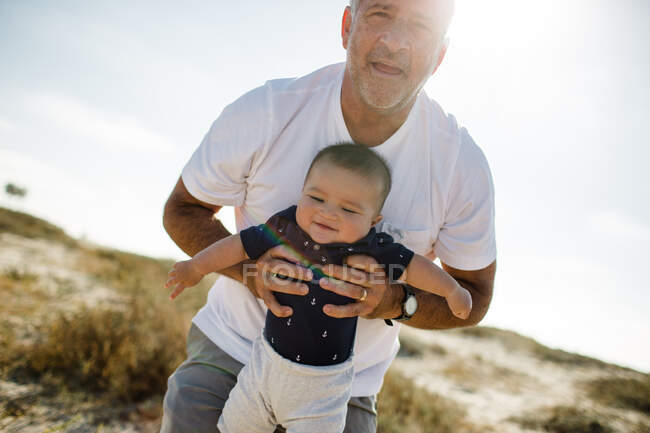 Abuelo sosteniendo & Jugando con su nieto mientras está de pie en la playa - foto de stock