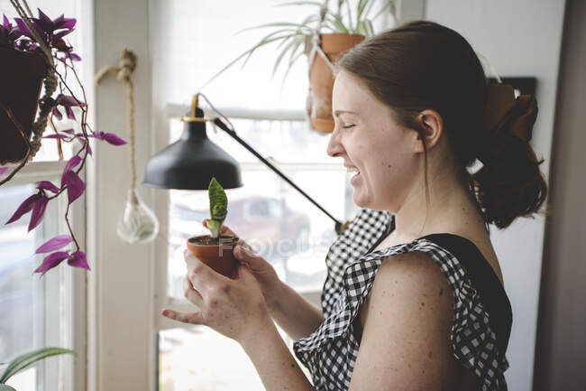 Mujer joven sonríe y se ríe de una de sus plantas en una habitación luminosa - foto de stock