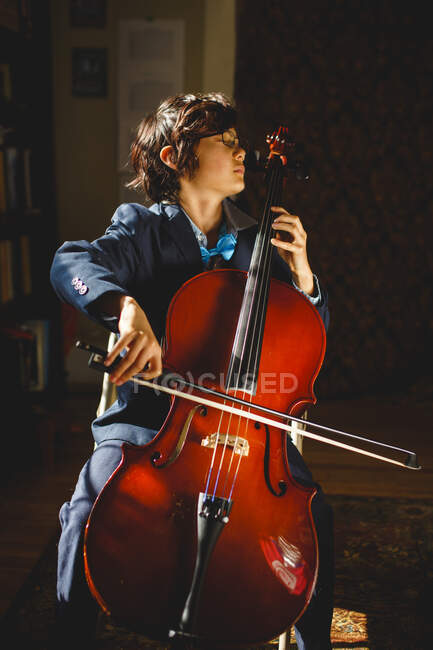 A serious boy in suit and bow tie with glasses plays cello at home — Stock Photo