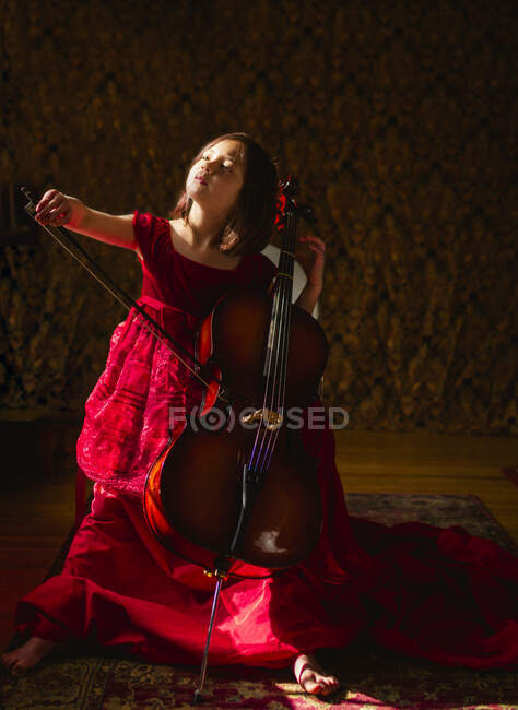 A small beautiful child in long red dress plays cello in window light — Stock Photo