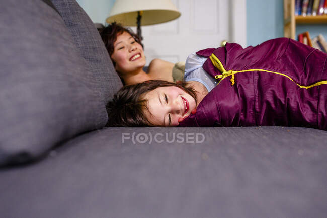 Dos niños sonrientes felices yacían juntos en un sofá en sacos de dormir - foto de stock