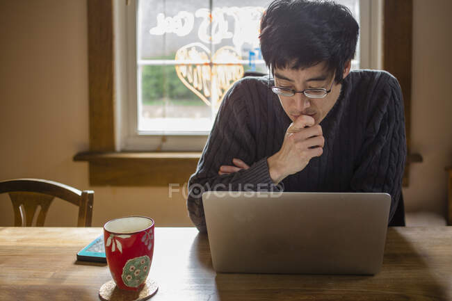 Un hombre se sienta en una mesa de comedor en la computadora trabajando desde casa - foto de stock