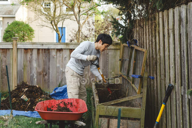 Un hombre usa una horquilla para palear compost en una carretilla roja - foto de stock