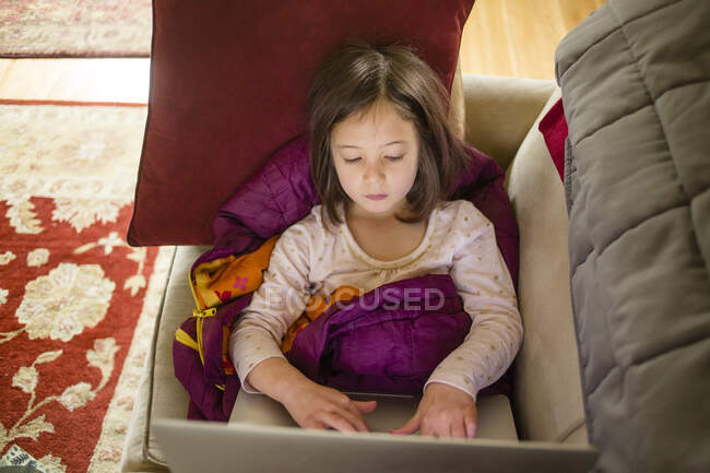 A little girl sits on couch in a pile of blankets working on computer — Stock Photo