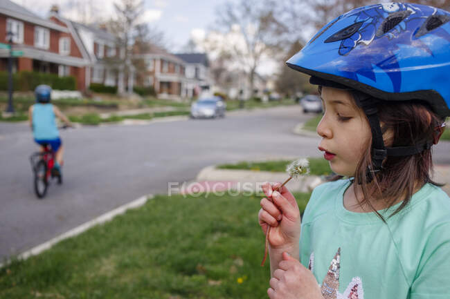 Маленькая девочка дует на одуванчик, в то время как мальчик катается на велосипеде по улице позади нее — стоковое фото