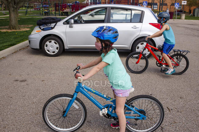 Un niño y una niña con máscaras en la cara montan bicicletas juntos en un estacionamiento - foto de stock