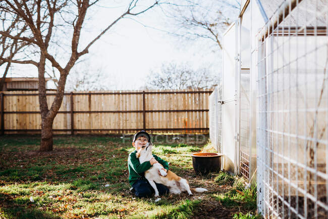 Young boy sitting outside backyard greenhouse hugging corgi dog — Stock Photo