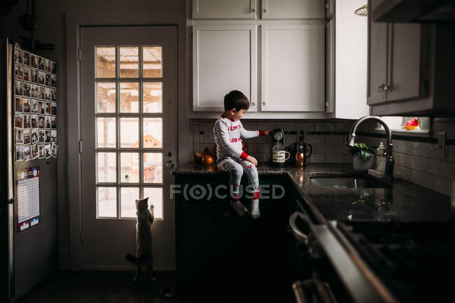 Kleiner Junge sitzt auf einem Tresen und kocht Kaffee mit Katze, die aus dem Fenster schaut — Stockfoto