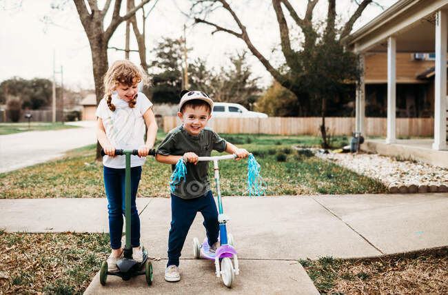 Младшие брат и сестра катаются на скутерах перед домом — стоковое фото