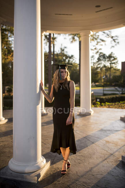Estudiante universitario en patio posando por una columna con gorra de graduación - foto de stock