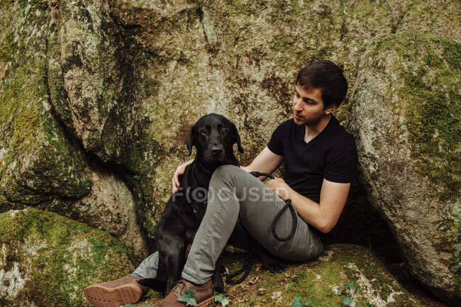 Jeune homme avec un labrador noir récupérateur assis sur un rocher mousseux — Photo de stock