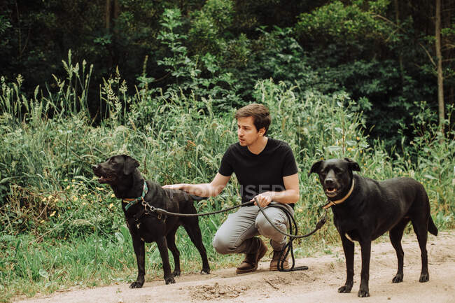 Millennial man sitting with his two black dogs on alert in the forest — Stock Photo