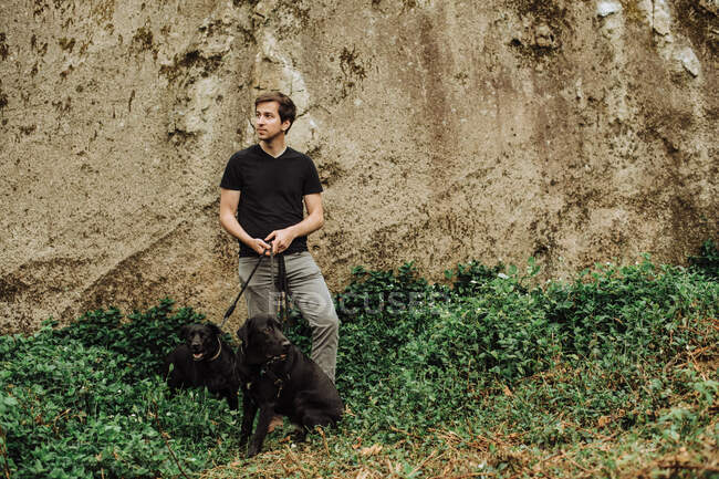Jeune homme avec deux chiens noirs en laisse debout contre le rocher — Photo de stock