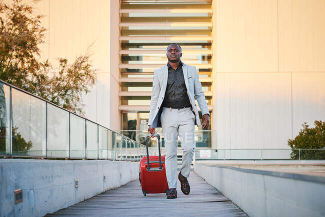 Uomo afro-americano con un abito elegante e una valigia rossa che cammina lungo una passerella di legno al tramonto. Uomo d'affari in viaggio. — Foto stock