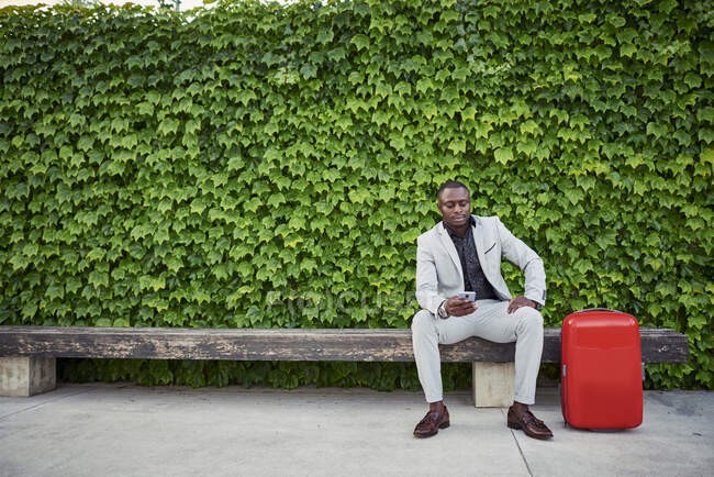 Empresario sentado en un banco con una maleta roja y un teléfono móvil en la mano. Hombre de negocios viajando. - foto de stock