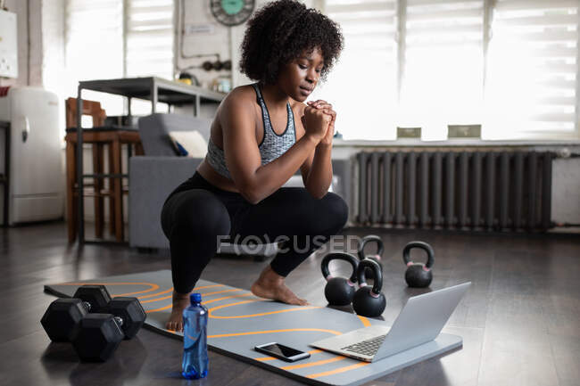 Чорна леді застібає руки і присідає біля ноутбука під час онлайн-тренувань вдома — стокове фото