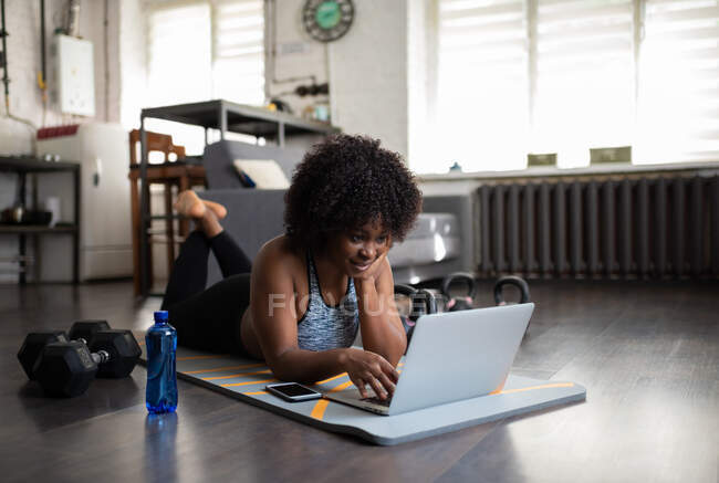 Erwachsene schwarze Athletin liegt beim Training zu Hause auf Matte und blättert im Laptop — Stockfoto