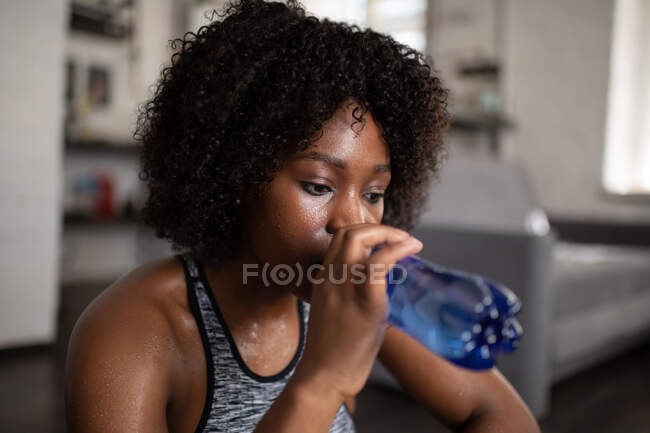 Femelle noire fatiguée sirotant l'eau de la bouteille pendant la pause dans l'entraînement à la maison — Photo de stock