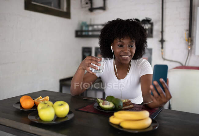 Mulher negra feliz com vidro de água fazendo chamada de vídeo para a família durante o café da manhã — Fotografia de Stock