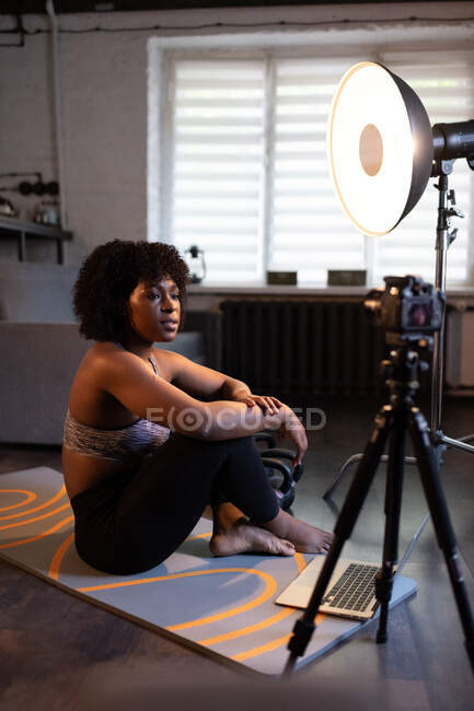 Черная женщина сидит на коврике и разговаривает с камерой во время записи онлайн тренировки — стоковое фото