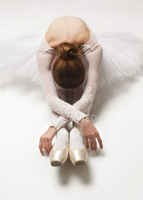 Приваблива балерина в білій балетній сукні та взутті, що сидить на підлозі, нахилена, студійний знімок — стокове фото