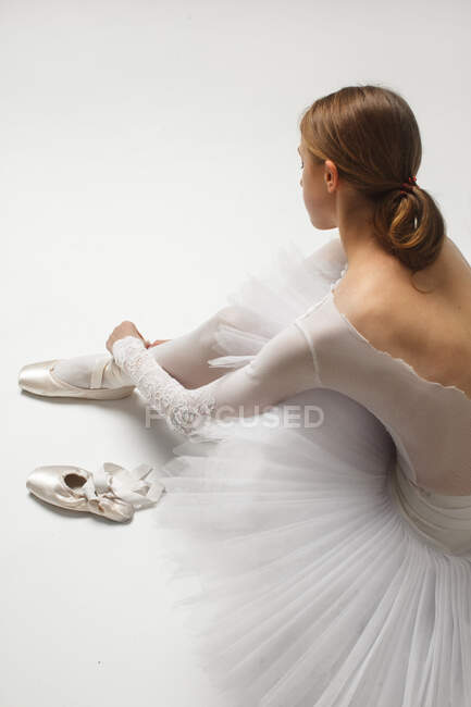 Балетна танцівниця зав'язує свої балетні туфлі навколо щиколотки на білій підлозі — стокове фото