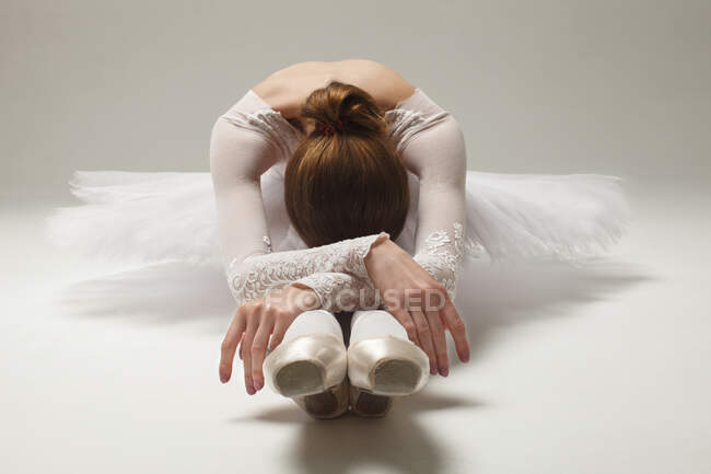 Красивая балерина в белой балетной одежде, сидящая на полу наклоненная, студийная съемка — стоковое фото