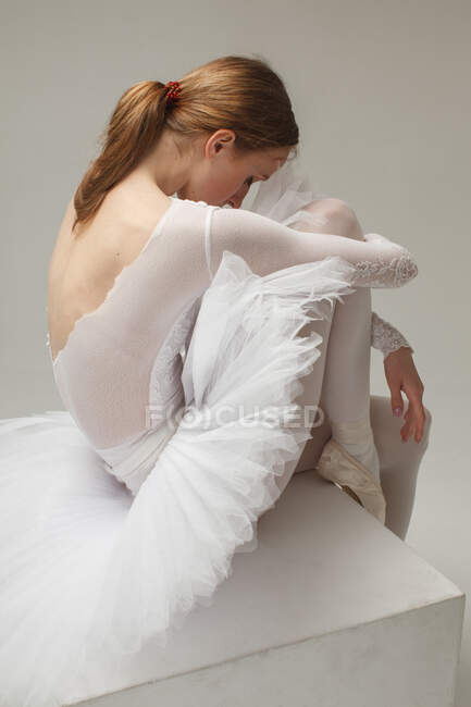 Чувственный портрет молодой балерины в белом платье, сидящей на кубе на фоне студии — стоковое фото