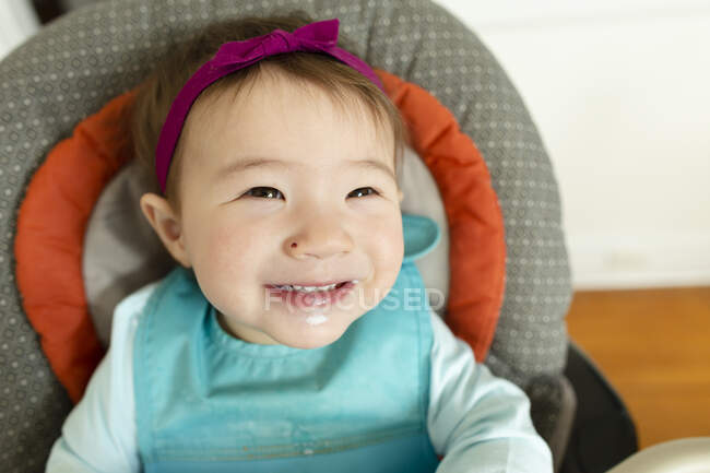 Liebenswert lächelndes Baby-Mädchen mit Stirnband sitzt im Hochstuhl zu Hause — Stockfoto