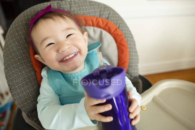 Sorrindo bebê menina segura xícara de leite enquanto sentado em cadeira alta em casa — Fotografia de Stock