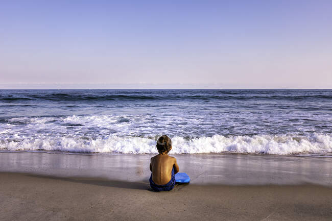 Visão traseira de criança de 5 anos sentada na praia de frente para o oceano — Fotografia de Stock