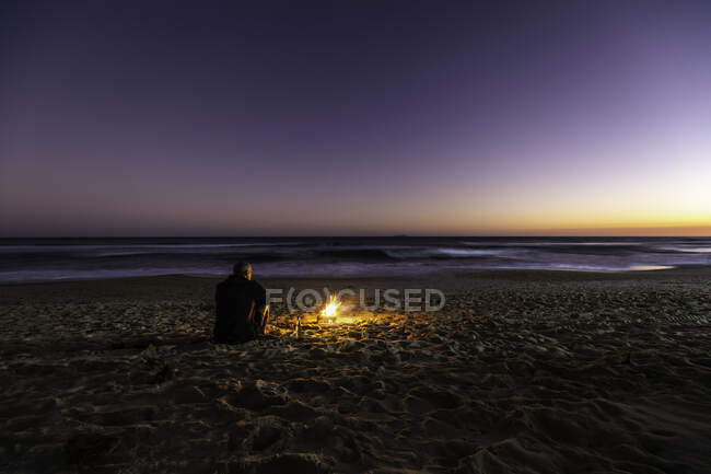 Вид сзади на одинокого парня и костер на пляже на закате — стоковое фото