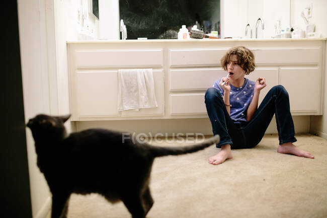 Adolescente chica se sienta en el suelo mientras se cepilla los dientes con gato cercano - foto de stock