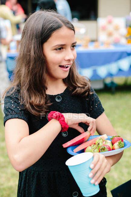 Tween niña sonríe en una celebración mientras lleva un plato de fruta - foto de stock