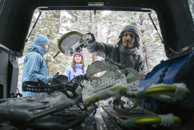 Joven hombre afroamericano levanta sus esquís nevados del maletero mientras sus amigos hablan - foto de stock