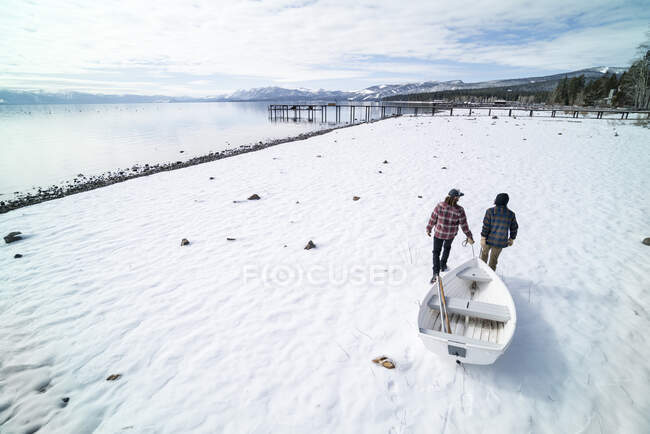 Two men pull a white rowboat across a snowy shore in South Lake Tahoe, CA — Stock Photo