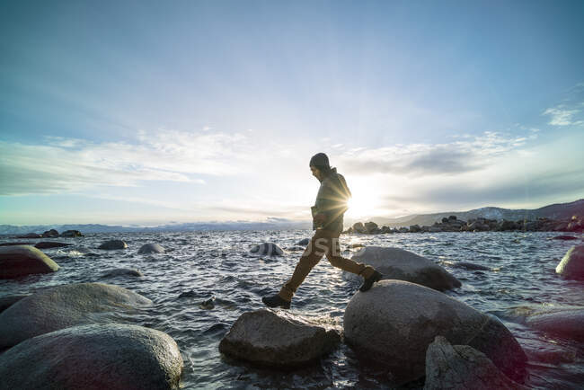 Homem segurando uma garrafa térmica salta de rocha em rocha através do Lago Tahoe — Fotografia de Stock