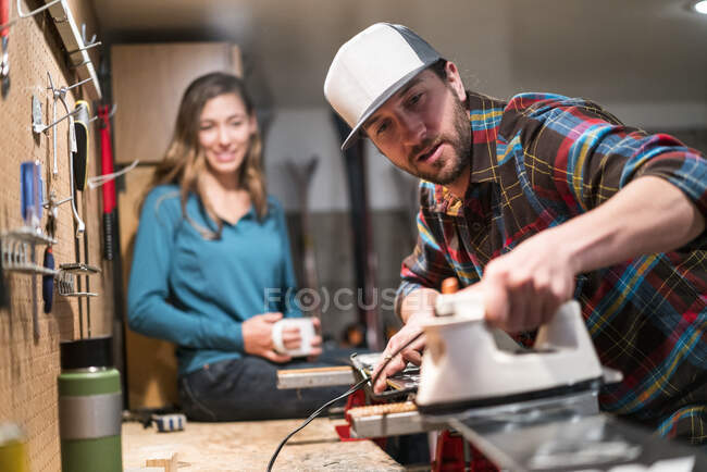 A man waxes his skis while a woman sits on the counter drinking tea in the background — Stock Photo