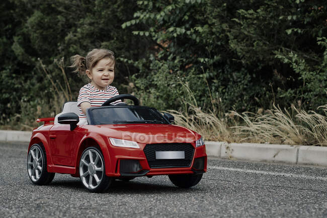 Una chica de 18 meses montada en un coche de juguete rojo - foto de stock