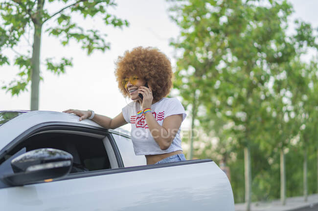 Mujer con el pelo afro hablando por teléfono al lado de su coche blanco - foto de stock