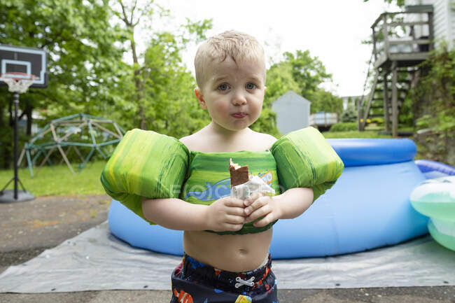 Young boy wearing swimming gear stands eating candy in yard with pool — Stock Photo