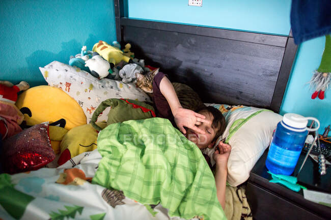 Мальчик в постели трёт глаза, когда просыпается с кошкой, делящей его подушку — стоковое фото