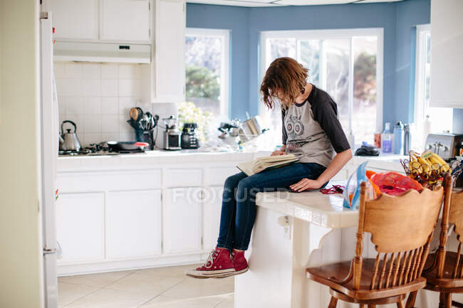 Chica adolescente se sienta en su encimera de cocina de azulejos leyendo un libro - foto de stock