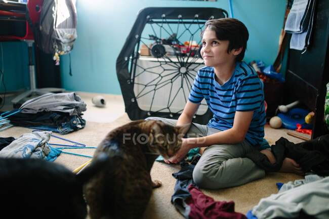 Niño de diez años se sienta distraído junto a su gato mientras hace la colada - foto de stock
