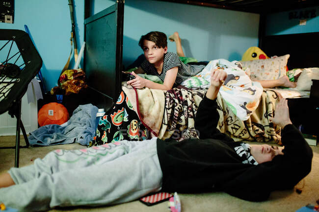 Мальчик на нижней койке играет в видеоигры, а сестра на полу — стоковое фото