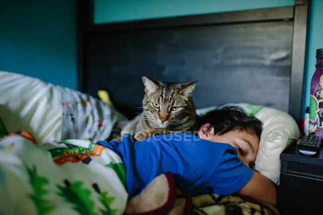 Chico despertando por la mañana mientras su gato agachado descansa sobre su espalda - foto de stock
