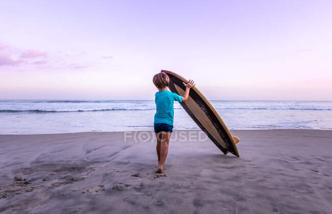 Rear view 4 years old kid carrying a wooden surfboard at the beach — Stock Photo