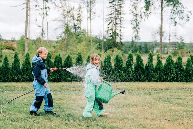 Brother and sister playing in the rain with a hose and watering can — Stock Photo