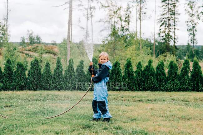 Ragazzo in piedi con un tubo spruzzando acqua a casa in cortile — Foto stock